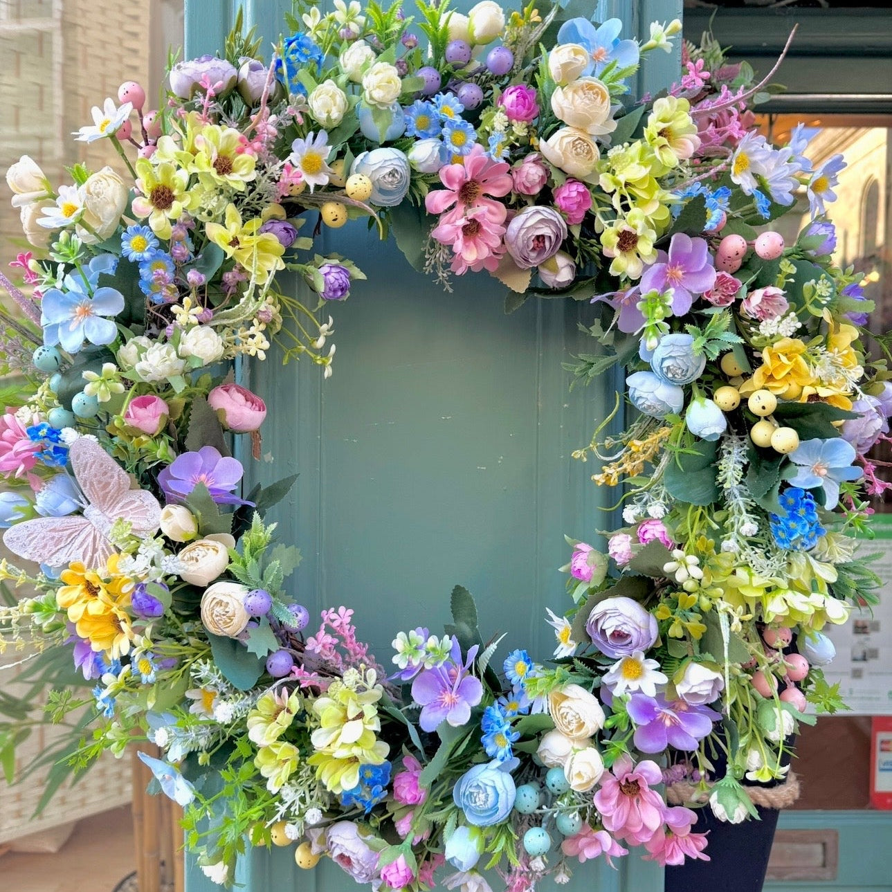 an artificial spring wreath hanging on a shop front, made with various spring pastel flower stems, like daisies, primroses and mini ranunculus 