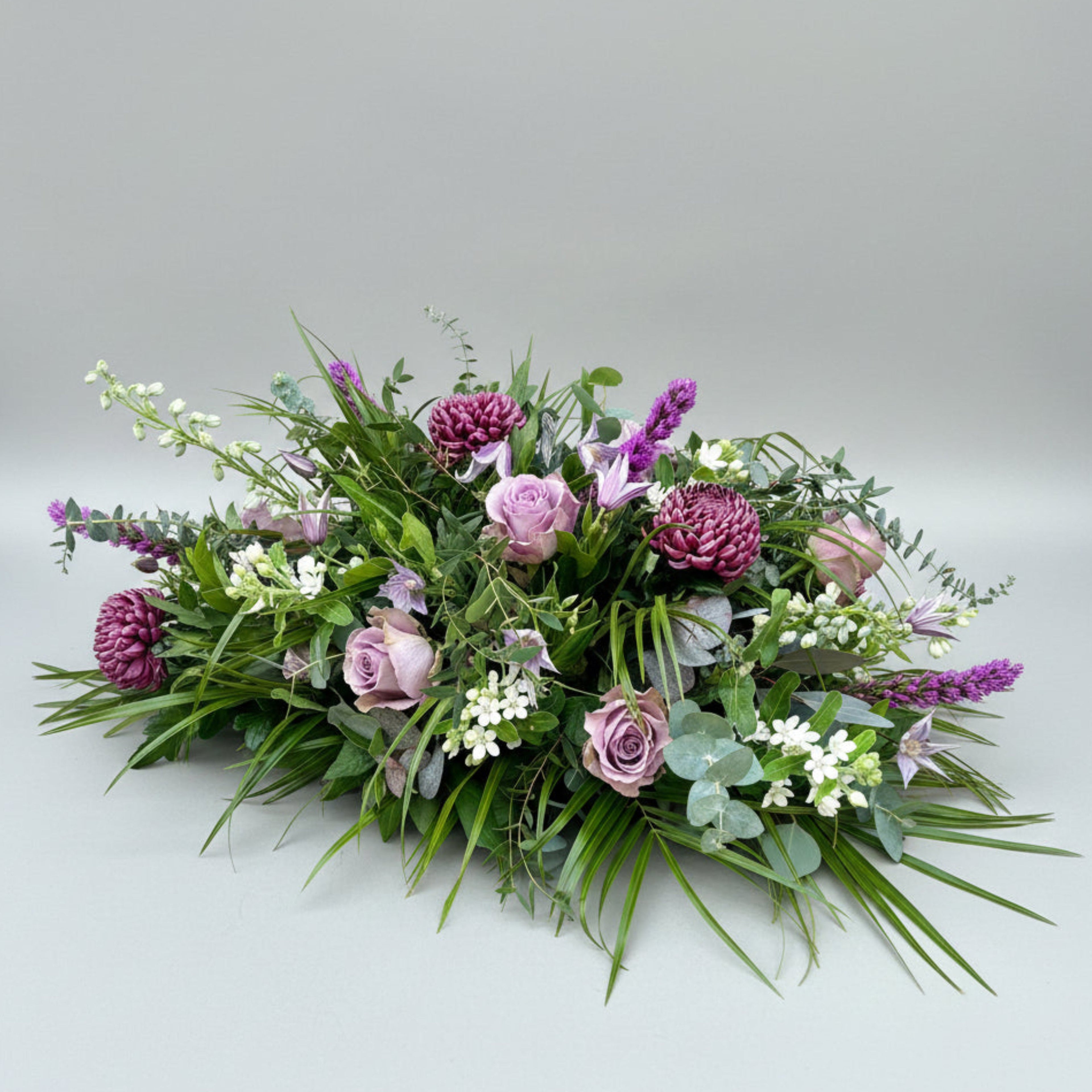 fresh flower funeral spray for casket with purple roses, chrysanthemums and clematis, white delphiniums and various foliage
