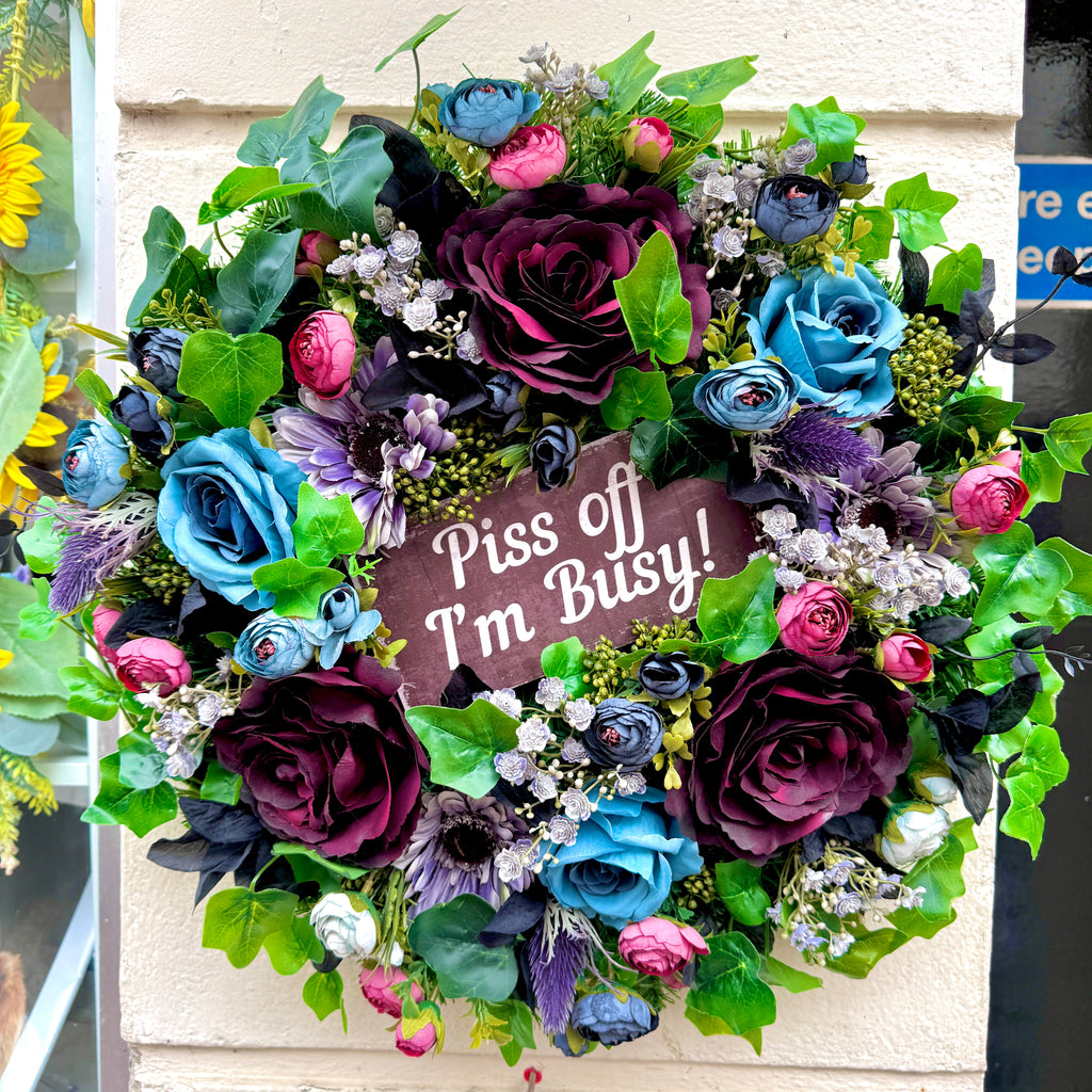 A wreath hanging on a wall with blue, burgundy and grey toned faux flowers with a piss off i'm busy sign