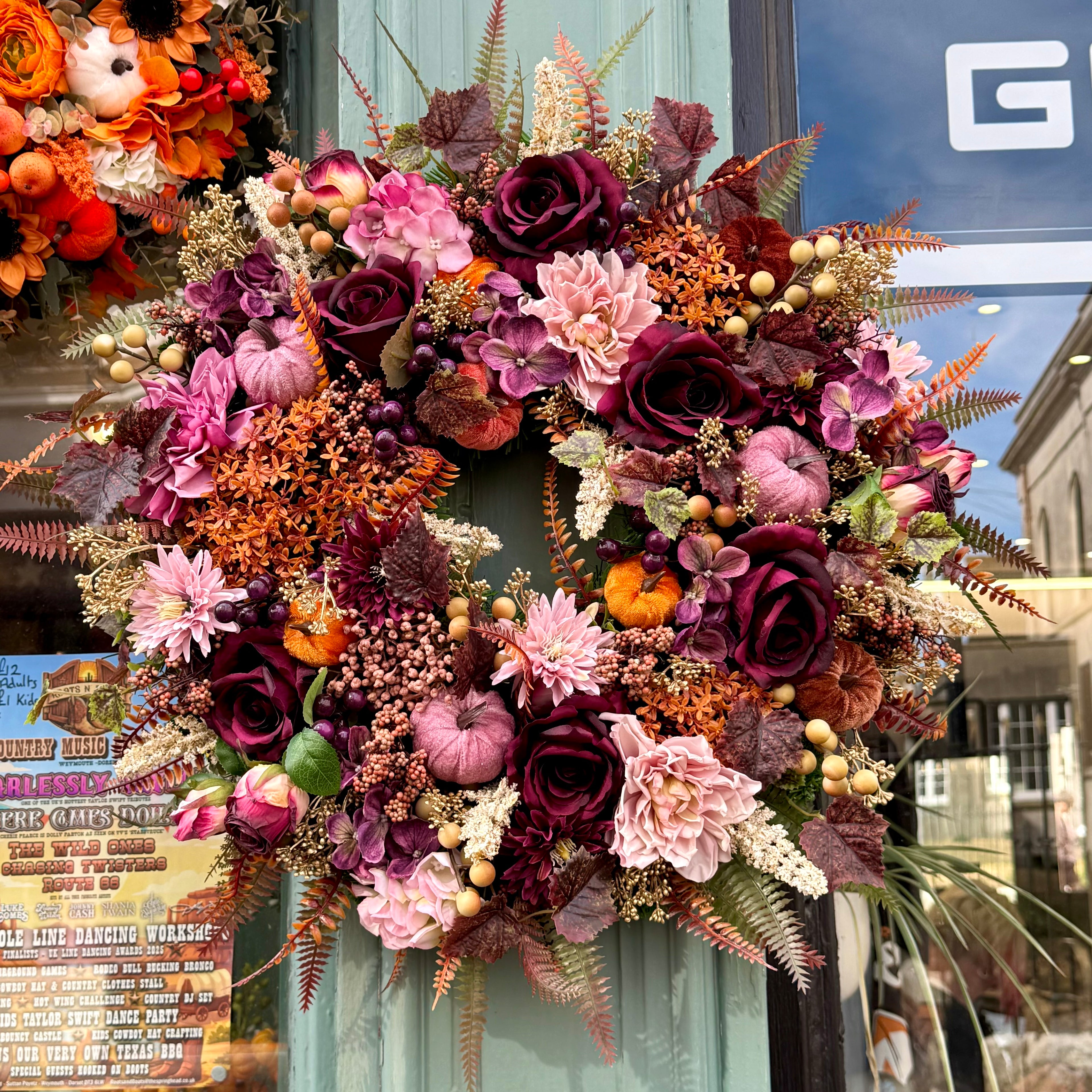 A large autumn-themed floral wreath hanging on a storefront window. The wreath features deep burgundy and pink roses, light pink chrysanthemums, small orange pumpkins, beige berries, and brown and green foliage.