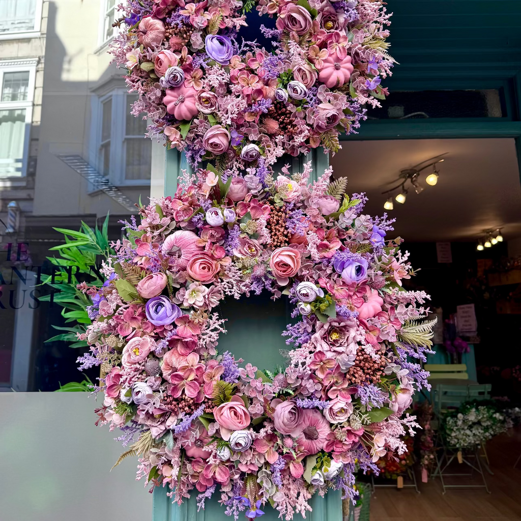 A large decorative floral wreath made of pink and purple faux flowers hangs vertically on a teal storefront door, with a shop interior and street scene visible in the background.