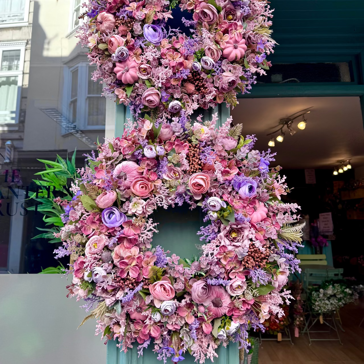 A large decorative floral wreath made of pink and purple faux flowers hangs vertically on a teal storefront door, with a shop interior and street scene visible in the background.