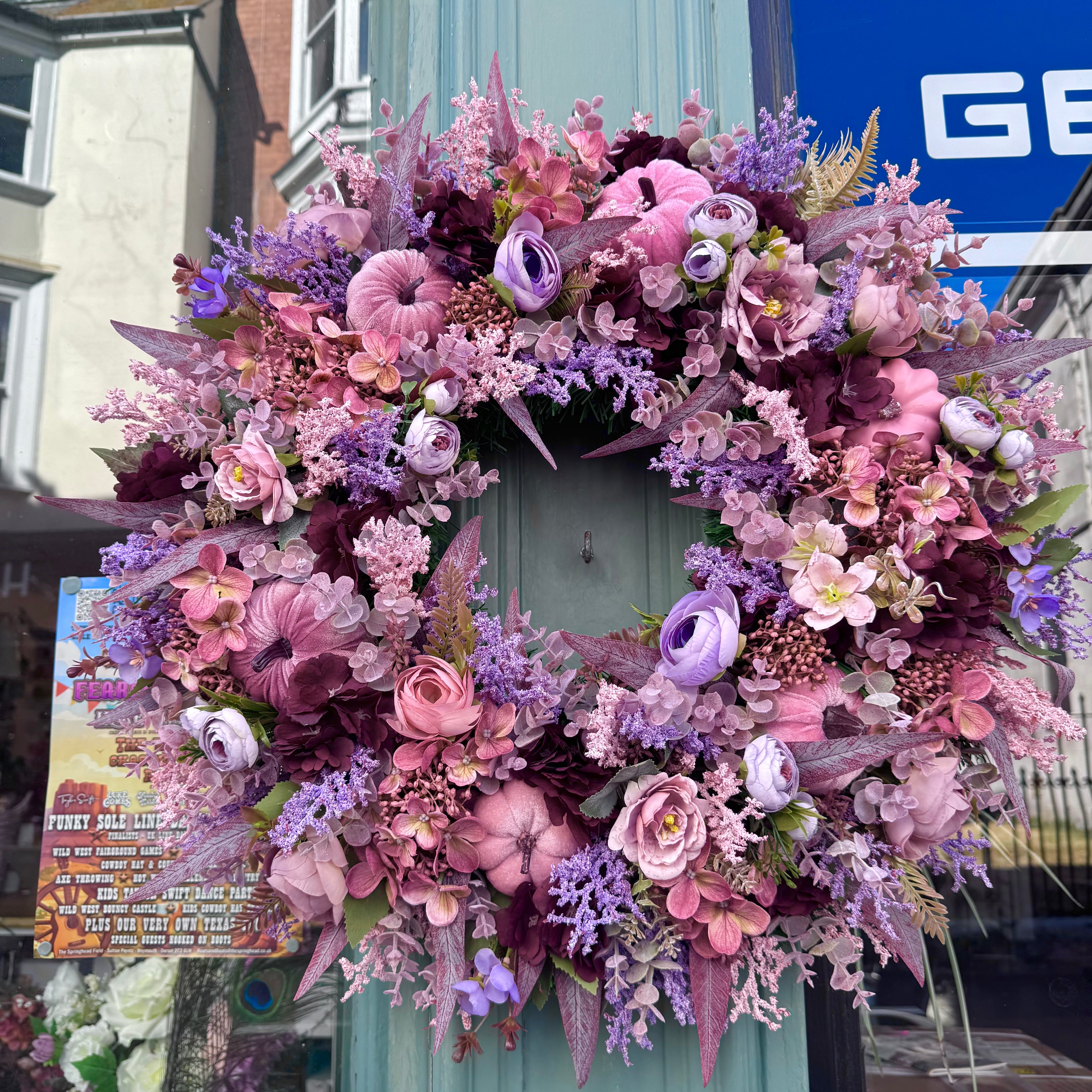 A large, lush artificial floral wreath in shades of pink, purple, and mauve, hanging on a teal-green wooden post outdoors in front of a storefront.