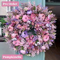 A large artificial floral wreath in shades of pink, purple, and lavender, decorated with faux pumpkins and dried botanicals, hanging on a teal door. A pink label in the bottom left corner reads 'Pumpkinella'.