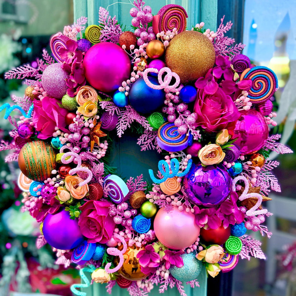 A vibrant, multicolored Christmas wreath decorated with pink and purple ornaments, candy-themed accents, and faux flowers, hanging on a teal door.