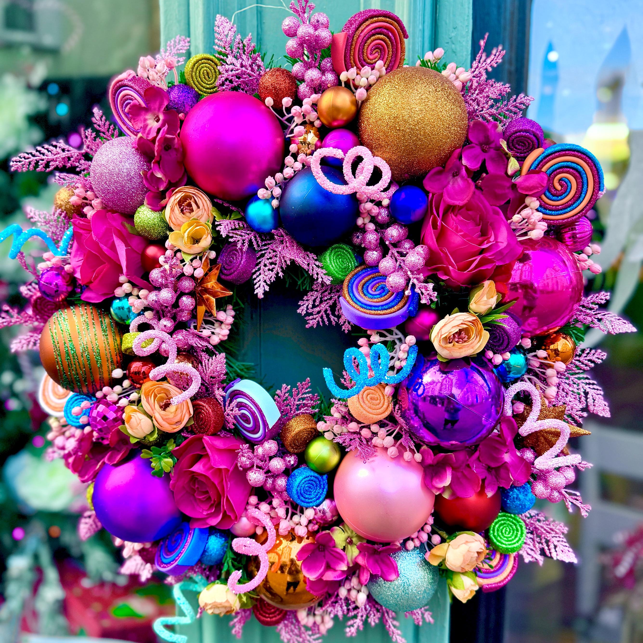 A vibrant, multicolored Christmas wreath decorated with pink and purple ornaments, candy-themed accents, and faux flowers, hanging on a teal door.