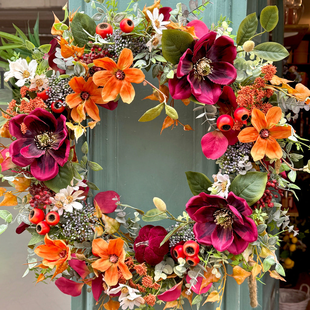 Colorful artificial, autumn floral wreath with orange and burgundy faux magnolias hanging on a teal shop front.