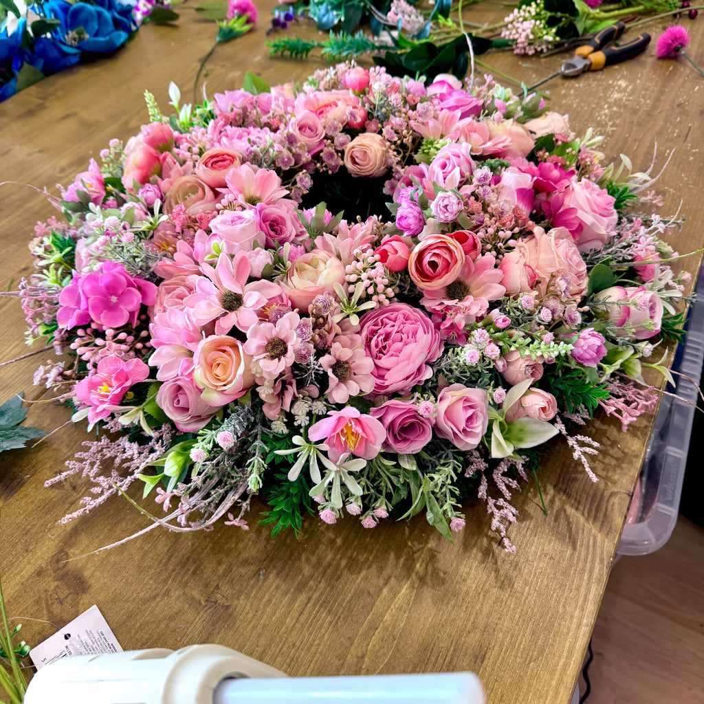 artificial pink wreath in the making on a wooden work bench with a glue gun and various flowers being cut
